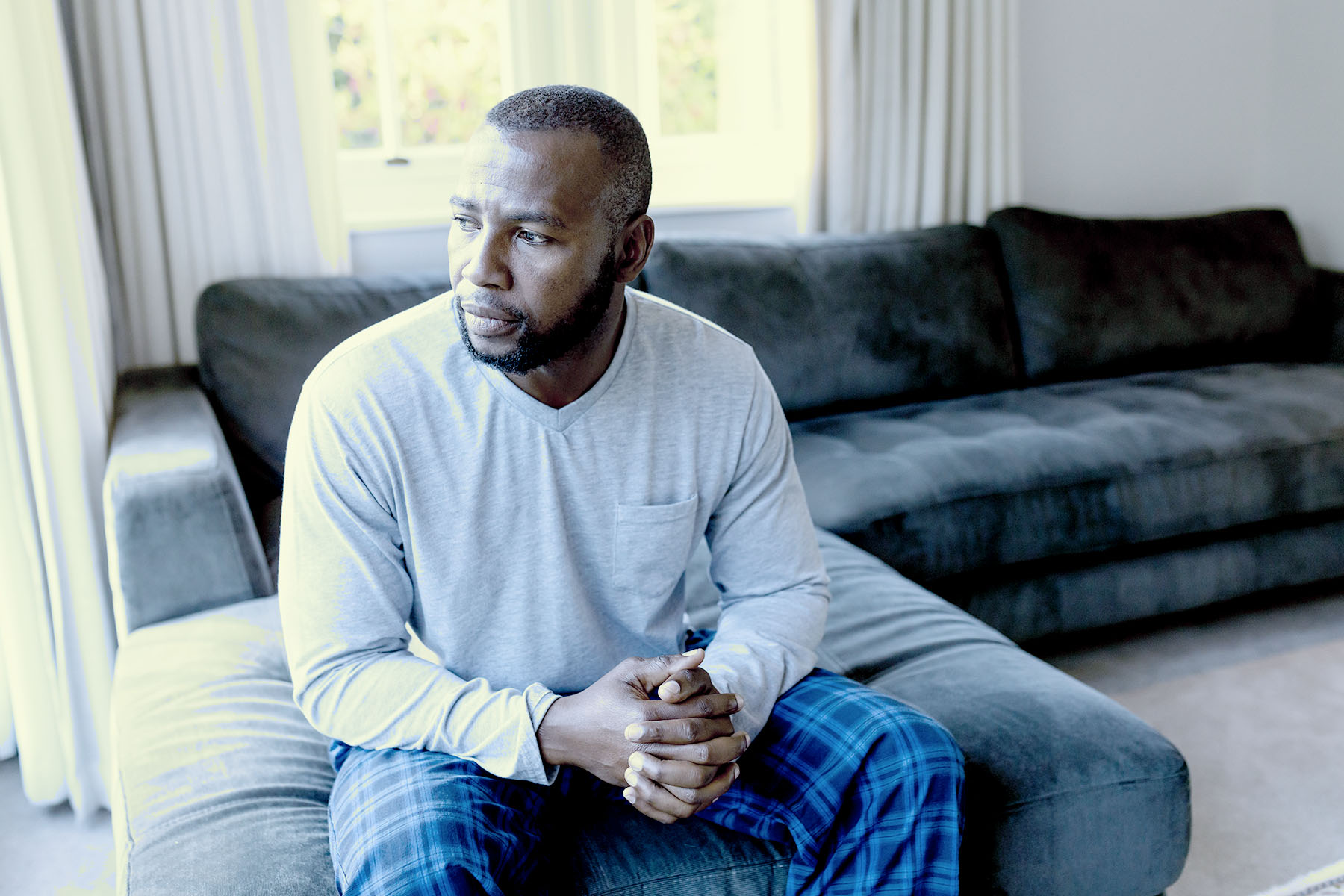 A man sitting on the edge of his couch in pajama bottoms and a sweatshirt, his hands clasped together as he looks pensively out a window.