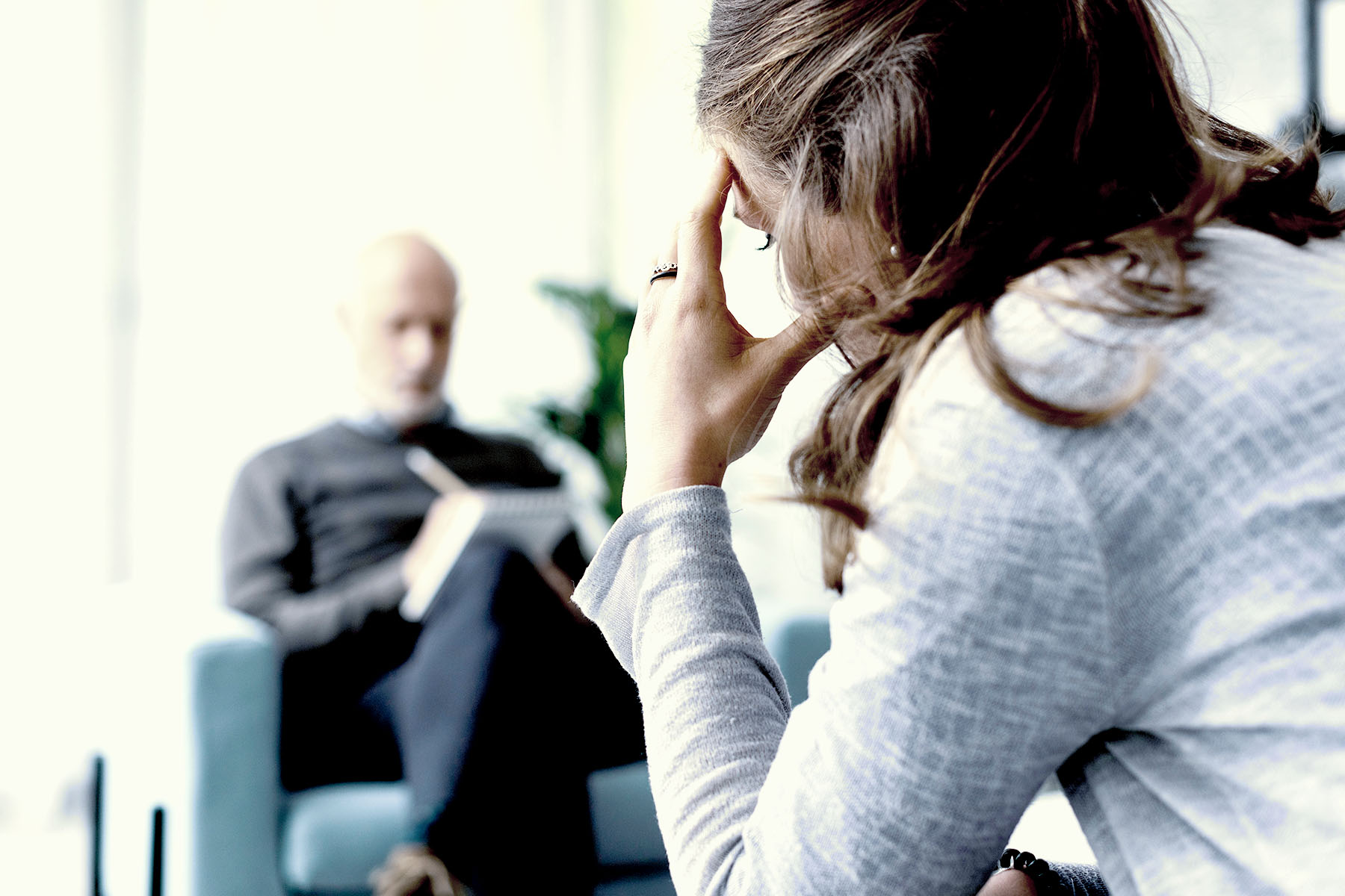 A man sitting on the edge of his couch in pajama bottoms and a sweatshirt, his hands clasped together as he looks pensively out a window.