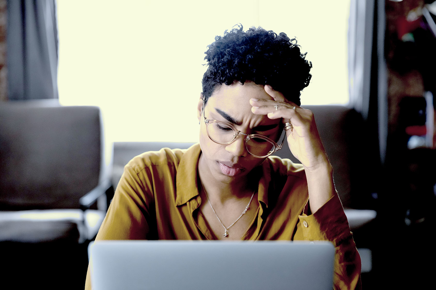 A man sitting on the edge of his couch in pajama bottoms and a sweatshirt, his hands clasped together as he looks pensively out a window.