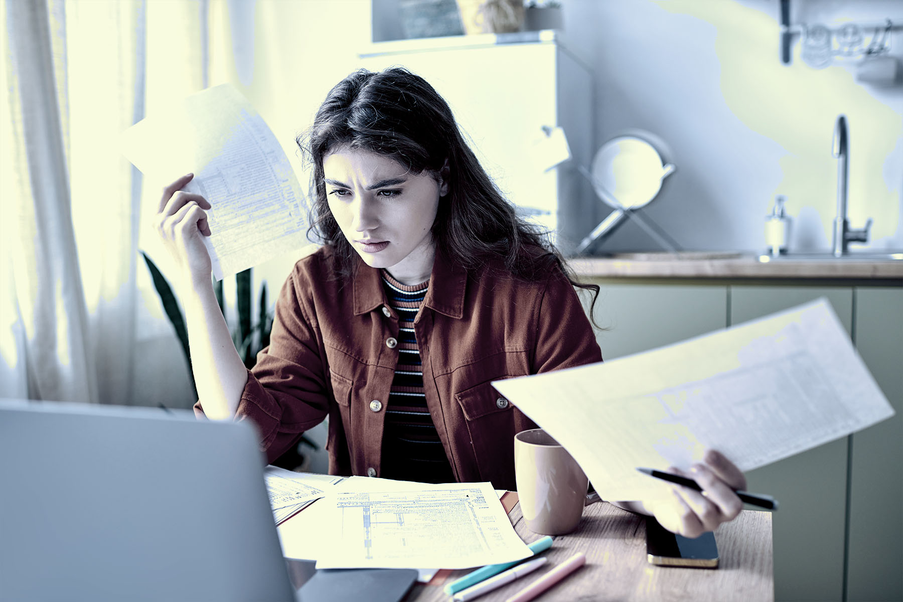 A young woman in front of a laptop, sorting through confusing forms at her desk, struggling to complete them.